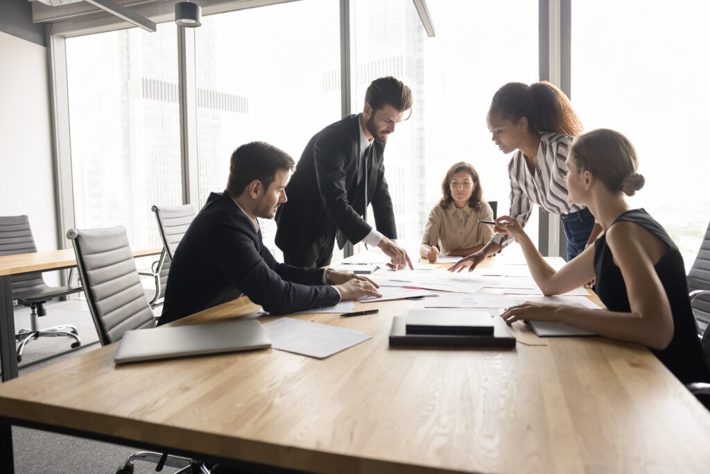 A group of business people in a meeting room standing at a table over paperwork in a discussion.