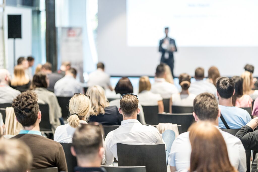 A person stands in front of a crowd on a stage at a conference.