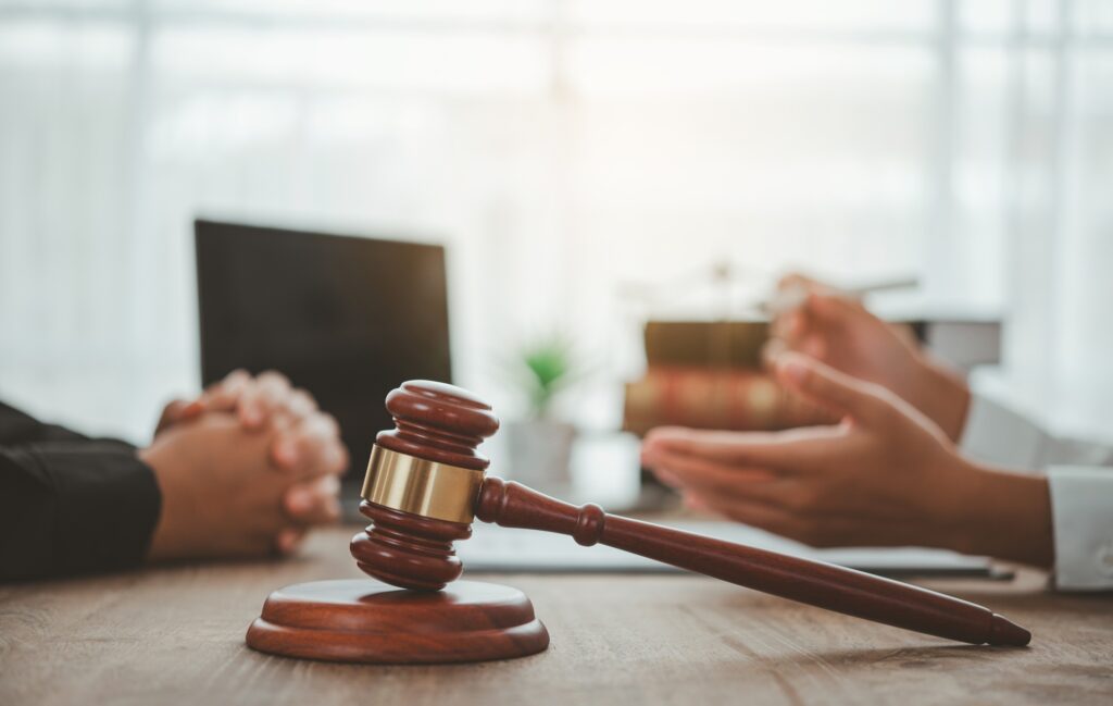 A lawyer sitting across a desk from a client next to a laptop and a gavel resting on its stand.