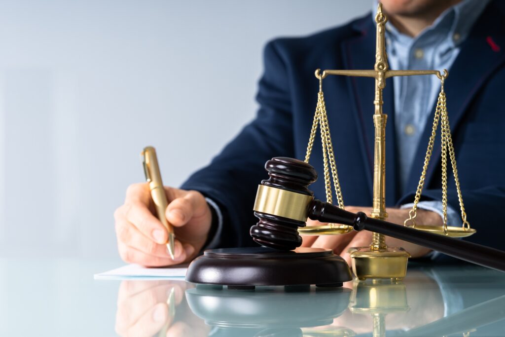 A male attorney writing on a piece of paper on a glass tabletop next to scales of justice and a gavel resting on its stand.