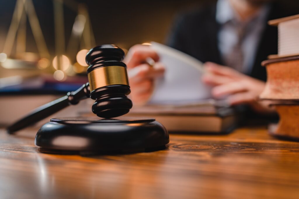 A gavel resting on its stand sitting on a wooden desk in front of an attorney looking at paperwork.