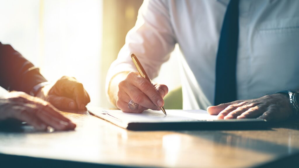 Two people sit at a desk in suits while one signs a piece of paper with a pen.