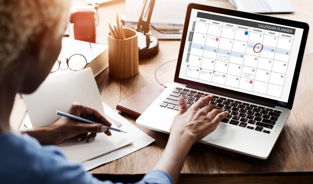 A person sits behind a silver laptop displaying a calendar on its screen with the 9th of the month circled in red.