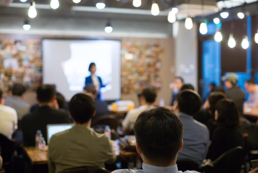 A speaker talks to a crowd in front of a screen while people listen in seats.